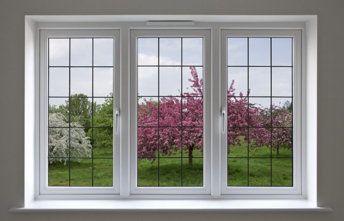 Beautifully made white wood framed windows with leaded glass panels, surrounded by a light grey wall. The view through the window is over an apple orchard under a cloudy blue sky. This is a composite of two images.