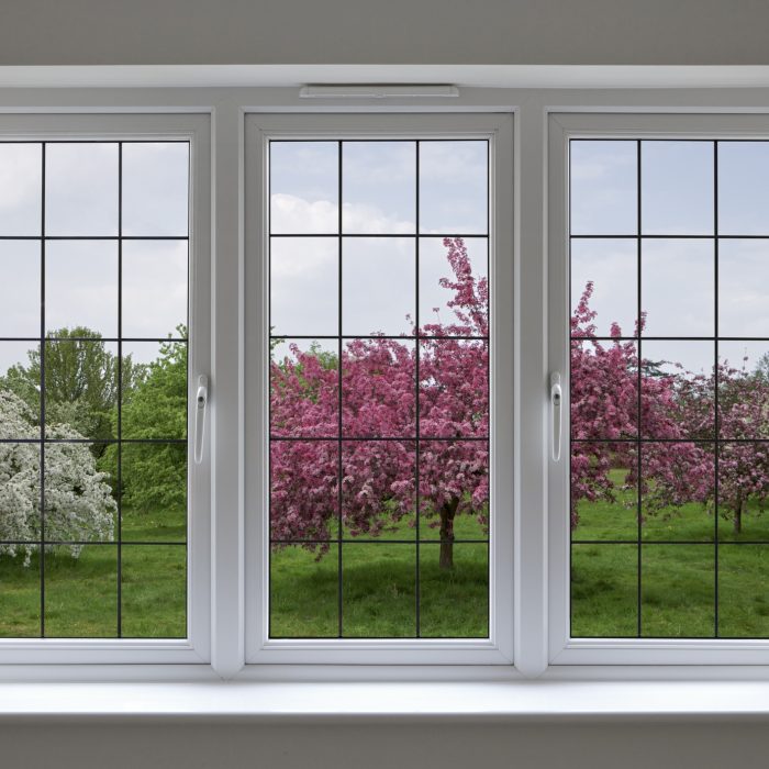 Beautifully made white wood framed windows with leaded glass panels, surrounded by a light grey wall. The view through the window is over an apple orchard under a cloudy blue sky. This is a composite of two images.